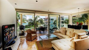 A spacious living room with a large sofa, a coffee table, and a television. There is a glass door leading to a balcony with palm trees and a view of a building. - Elements 217 by Tripintravel (Playa del Carmen)