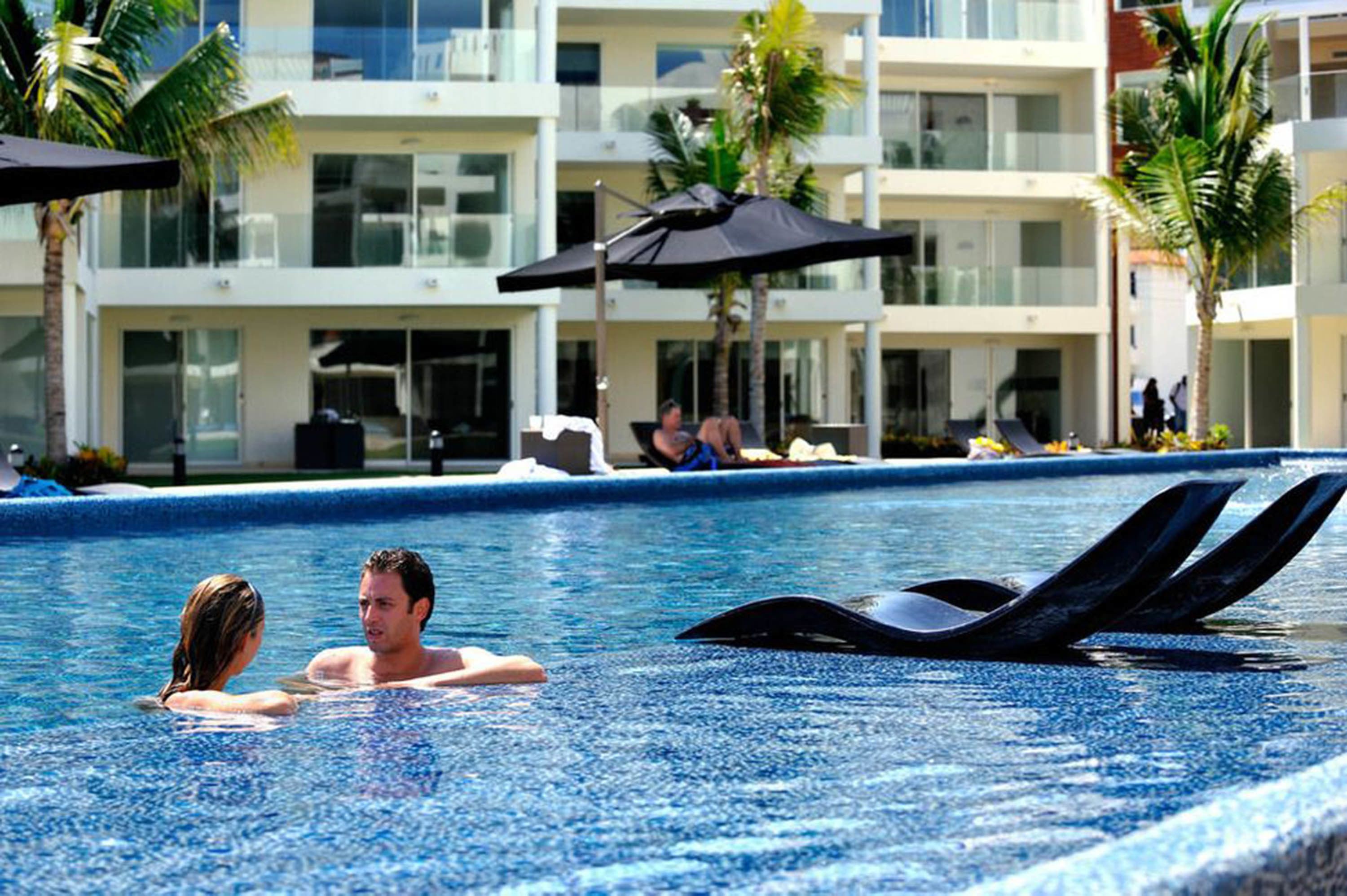 A swimming pool with lounge chairs and umbrellas, surrounded by palm trees and a modern building with balconies.