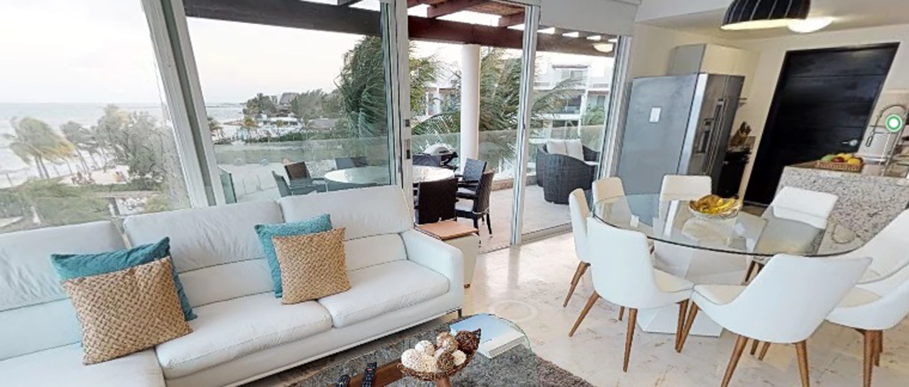 A modern living room with a glass dining table and white chairs, a white sofa with patterned cushions, and a view of the beach through large windows.