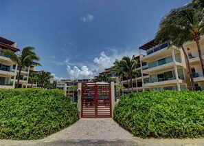 A resort entrance with a central gate, surrounded by lush greenery and palm trees, leading to multi-story buildings with balconies. - Elements GH09 by Tripintravel (Playa del Carmen)