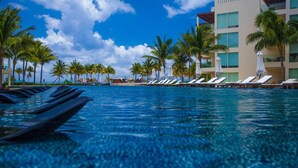 A pool with lounge chairs and palm trees, surrounded by a building with multiple floors and windows.