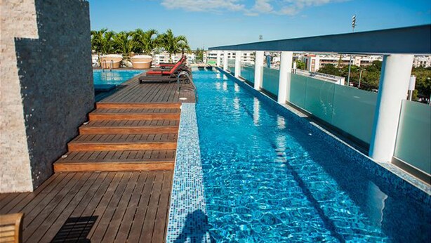 A rooftop infinity pool with wooden deck and lounge chairs, surrounded by a white railing and palm trees.