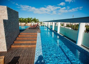 Une piscine à débordement située sur le toit, avec une terrasse en bois et des chaises longues, entourée d’une balustrade blanche et de palmiers.
