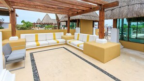 A covered outdoor seating area with a wooden pergola, a beige sofa with yellow accents, and a thatched-roof structure in the background. - Pueblito Luxury Penthouse by Tripintravel (Playa del Carmen)