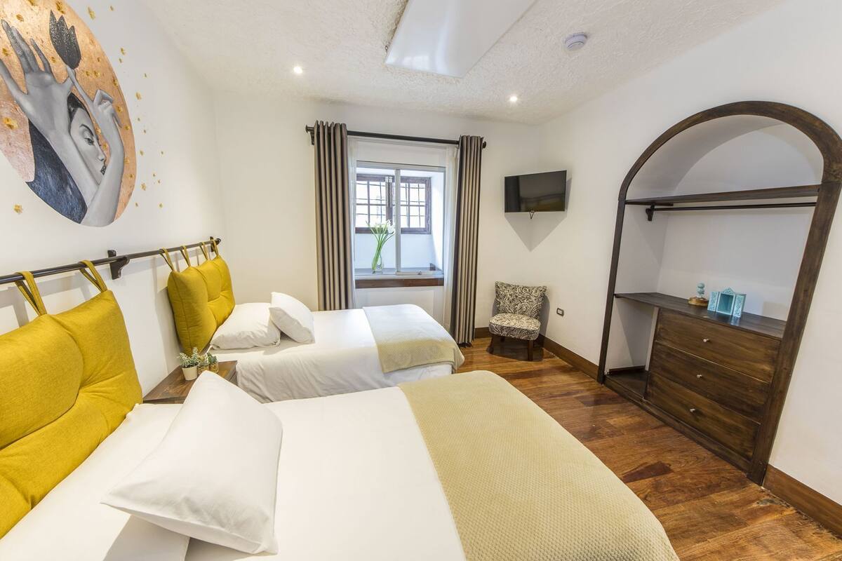 A hotel room with two beds, a chair, a dresser, a TV, and a window with curtains.