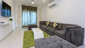 Ein modernes Wohnzimmer mit grauem Sofa, weißem Couchtisch und grauem Hocker. An der Wand ist ein Flachbildfernseher angebracht, es gibt einen Deckenventilator und eine Klimaanlage.