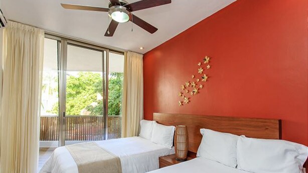 Habitación de hotel con dos camas, ventilador de techo y balcón con cortinas.