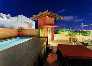 A rooftop pool area with a wooden deck, a small table with chairs, and a view of the city skyline at night.
