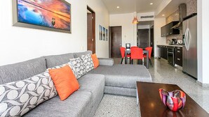 A modern living room with a grey sectional sofa, orange cushions, and a colorful painting. There is a dining area with red chairs and a kitchen with a stainless steel refrigerator. - Infinito 03 by Tripintravel (Playa del Carmen)