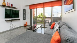 A modern living room with a flat-screen TV, a sofa with orange cushions, a coffee table, and a large window with a red blind. - Infinito 03 by Tripintravel (Playa del Carmen)