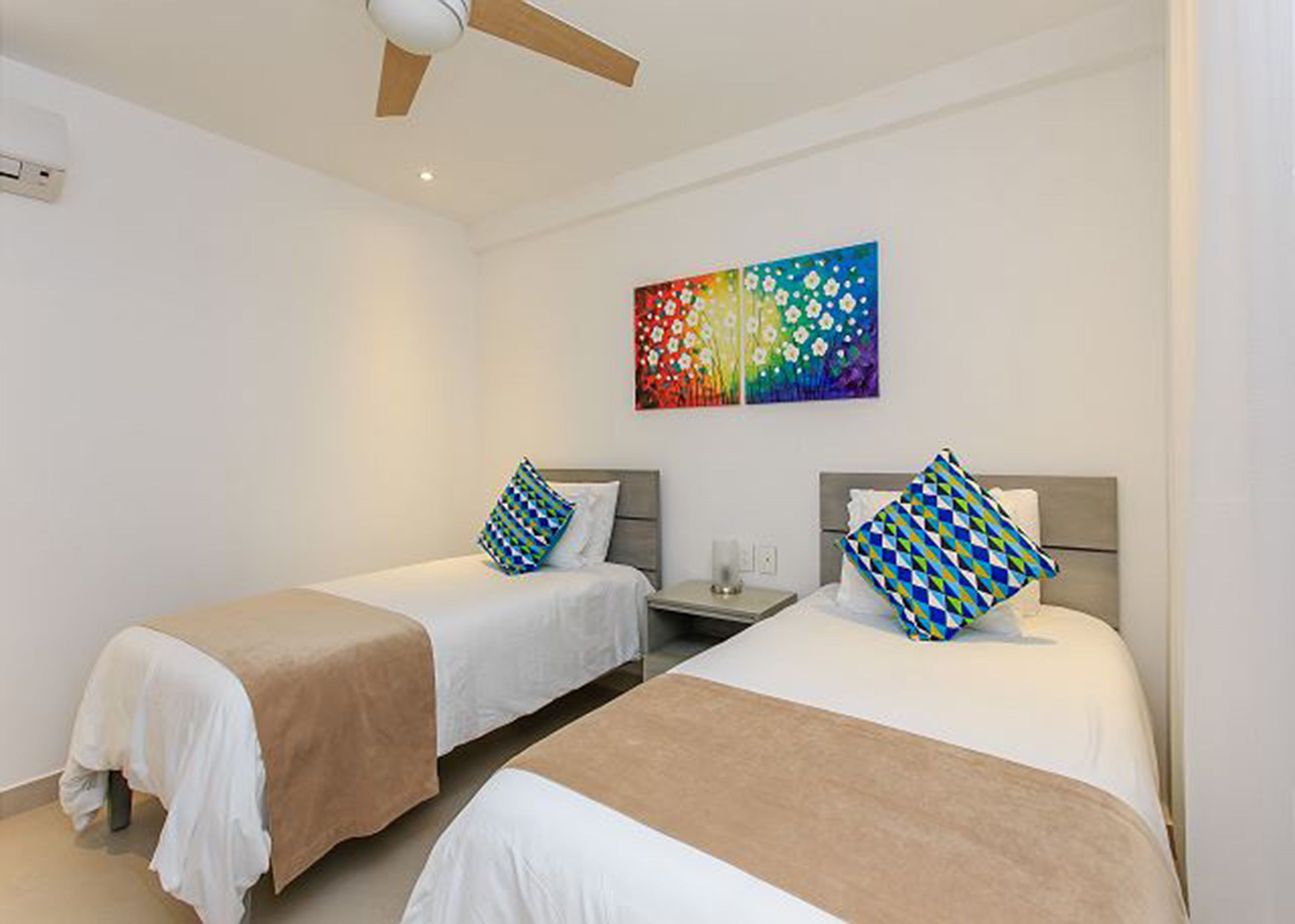 Une chambre d’hôtel avec deux lits, chacun agrémenté d’un oreiller à motifs, d’une table de chevet et d’un ventilateur de plafond.