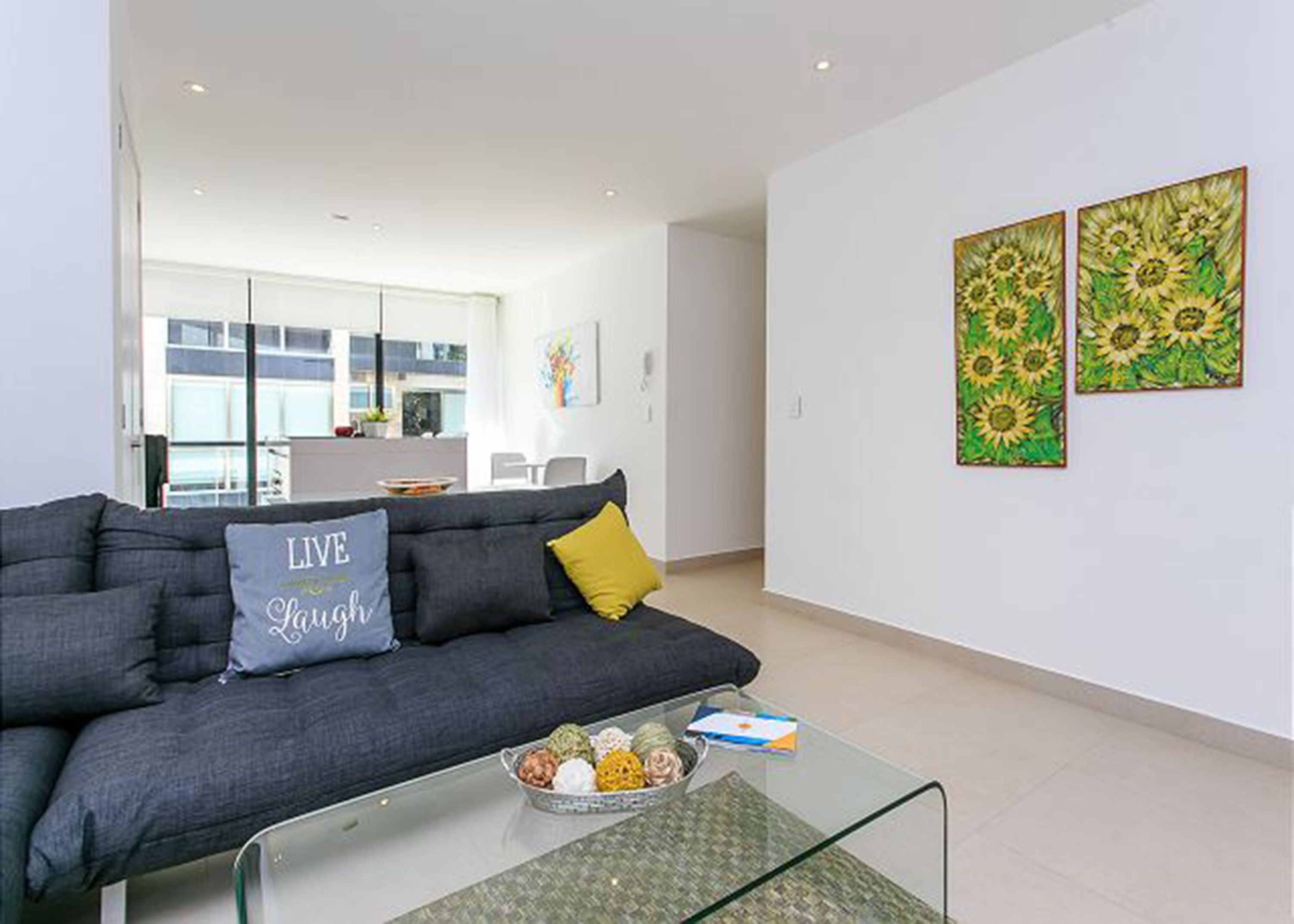 Ein modernes Wohnzimmer mit einem grauen Sofa, einem Glastisch und zwei Sonnenblumengemälden an der Wand.
