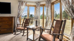 Una habitación con un aparador de madera, una mesa con lámpara, dos mecedoras y ventanas grandes con cortinas.