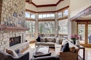 A cozy living room with a stone fireplace, large windows offering a view of a forested area, and a comfortable seating area.