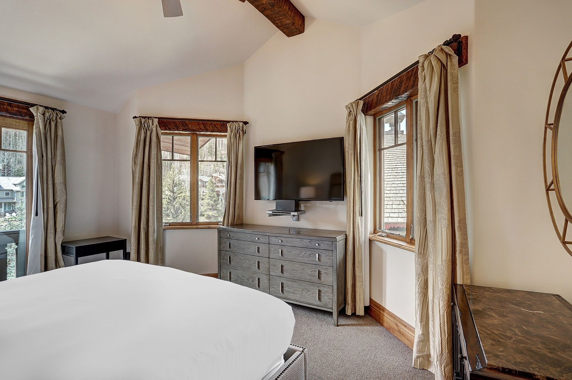 A bedroom with a bed, a dresser, a TV mounted on the wall, and large windows with curtains.