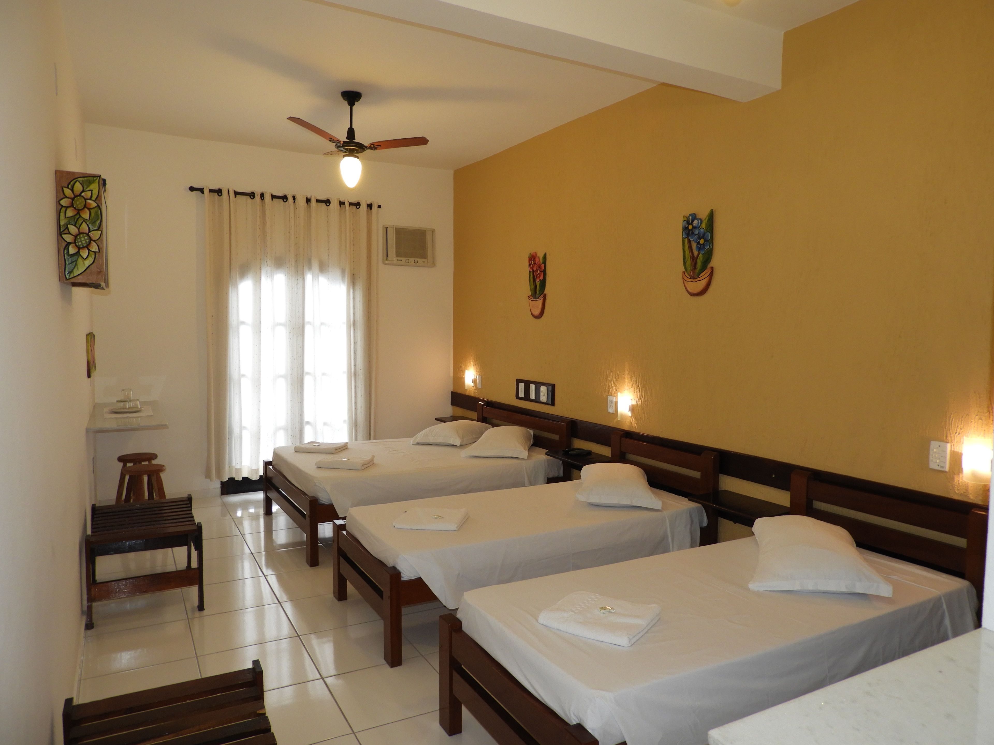 A hotel room with four single beds, a ceiling fan, a small table with a chair, and a window with curtains.