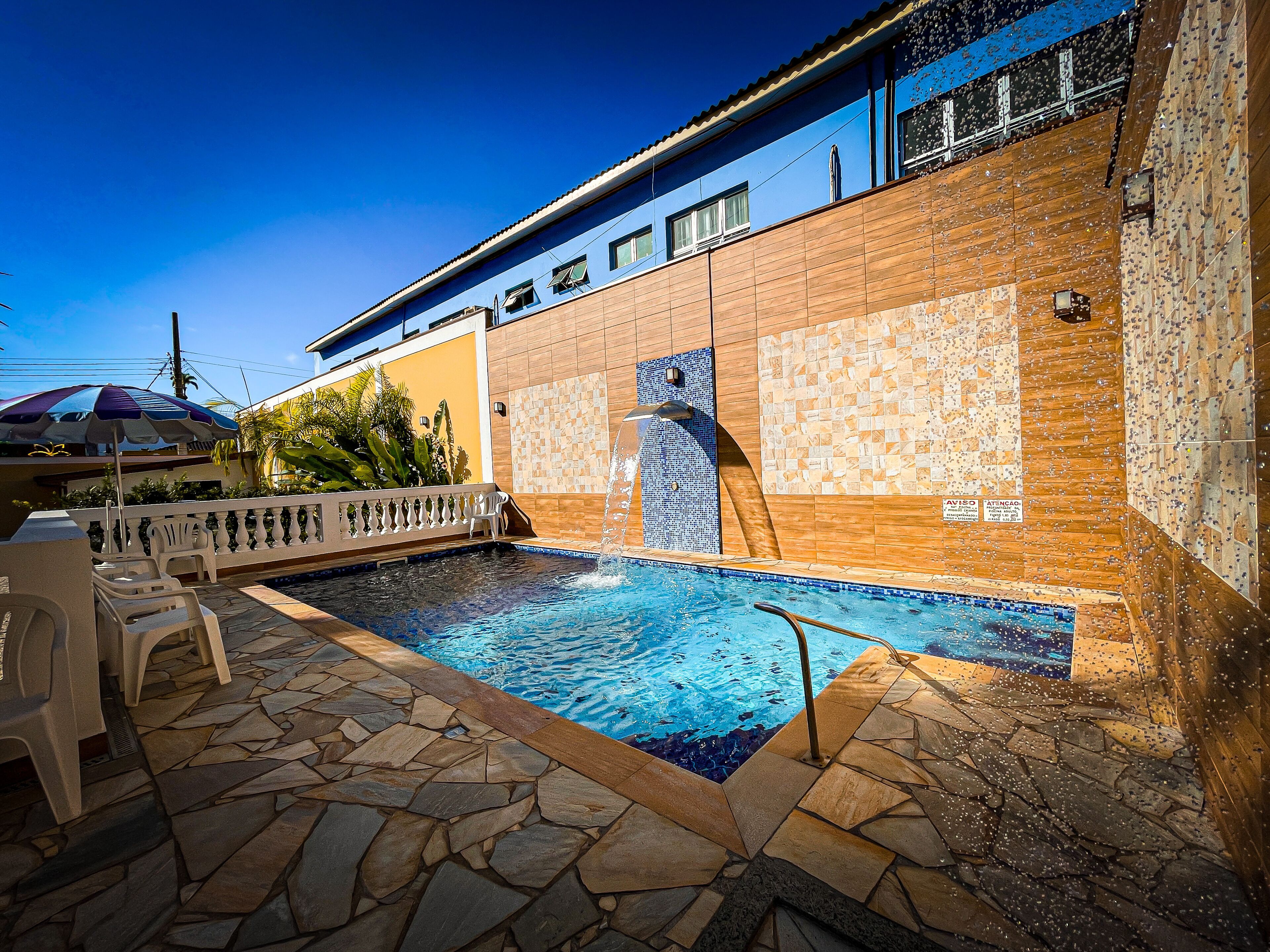 A swimming pool with stone tiles, a white fence, and a blue building in the background.