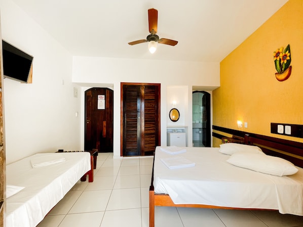 Habitación de hotel con dos camas individuales, ventilador de techo, un refrigerador pequeño y un baño visible a través de una puerta abierta.