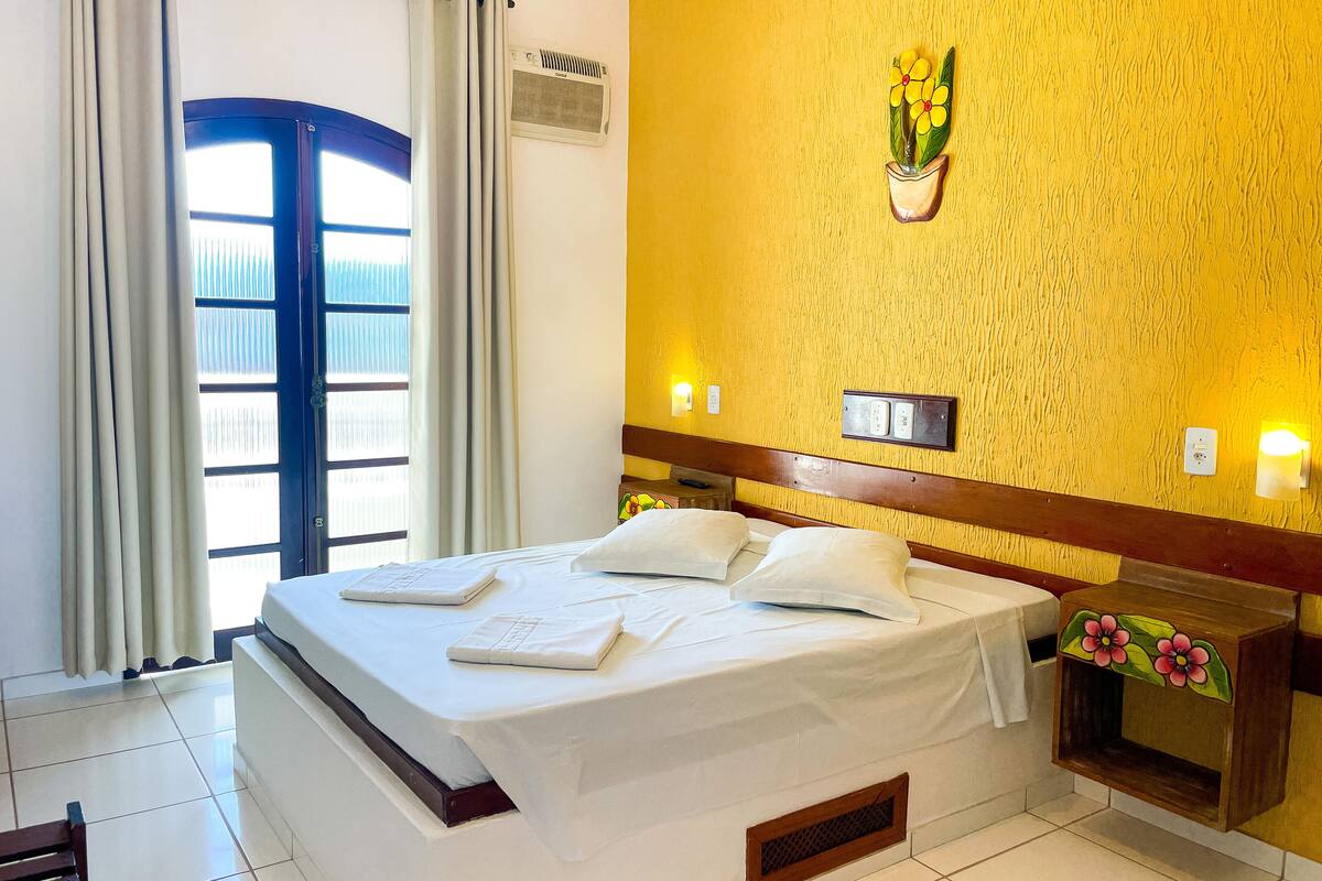 Habitación de hotel con cama, mesita de noche, aire acondicionado de pared, planta en maceta y ventana con cortinas.