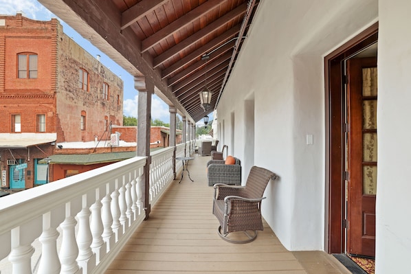 Une véranda couverte avec une balustrade blanche, des meubles en osier et une vue sur un bâtiment en briques rouges.