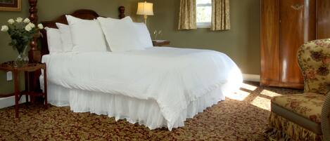 A neatly made bed with white linens, a wooden nightstand with a vase of flowers, a wooden wardrobe, and a floral-patterned armchair.