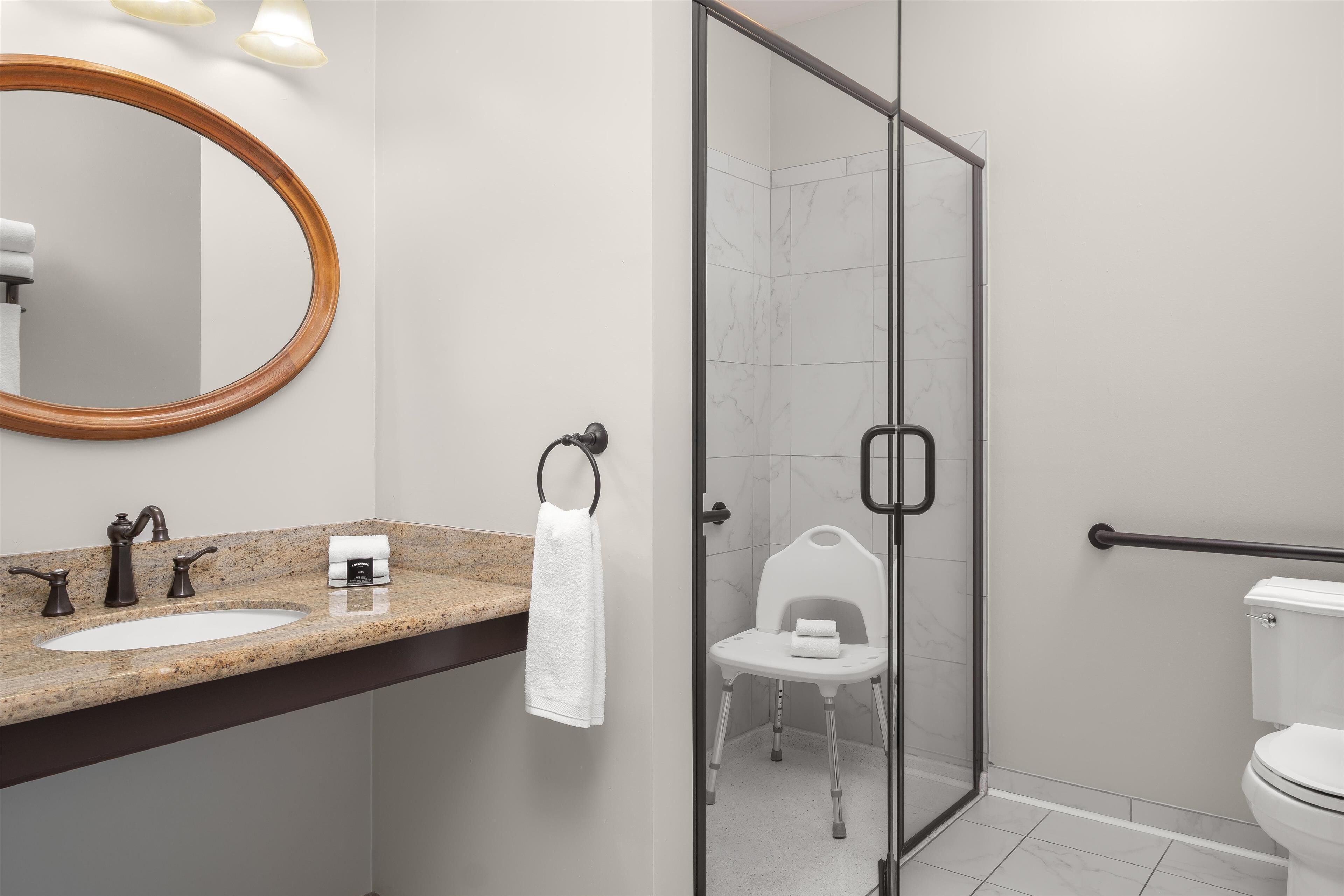 Et moderne badeværelse med en vask med granitbordplade, et rundt spejl, en glasafskærmet brusekabine med en hvid stol og et toilet.