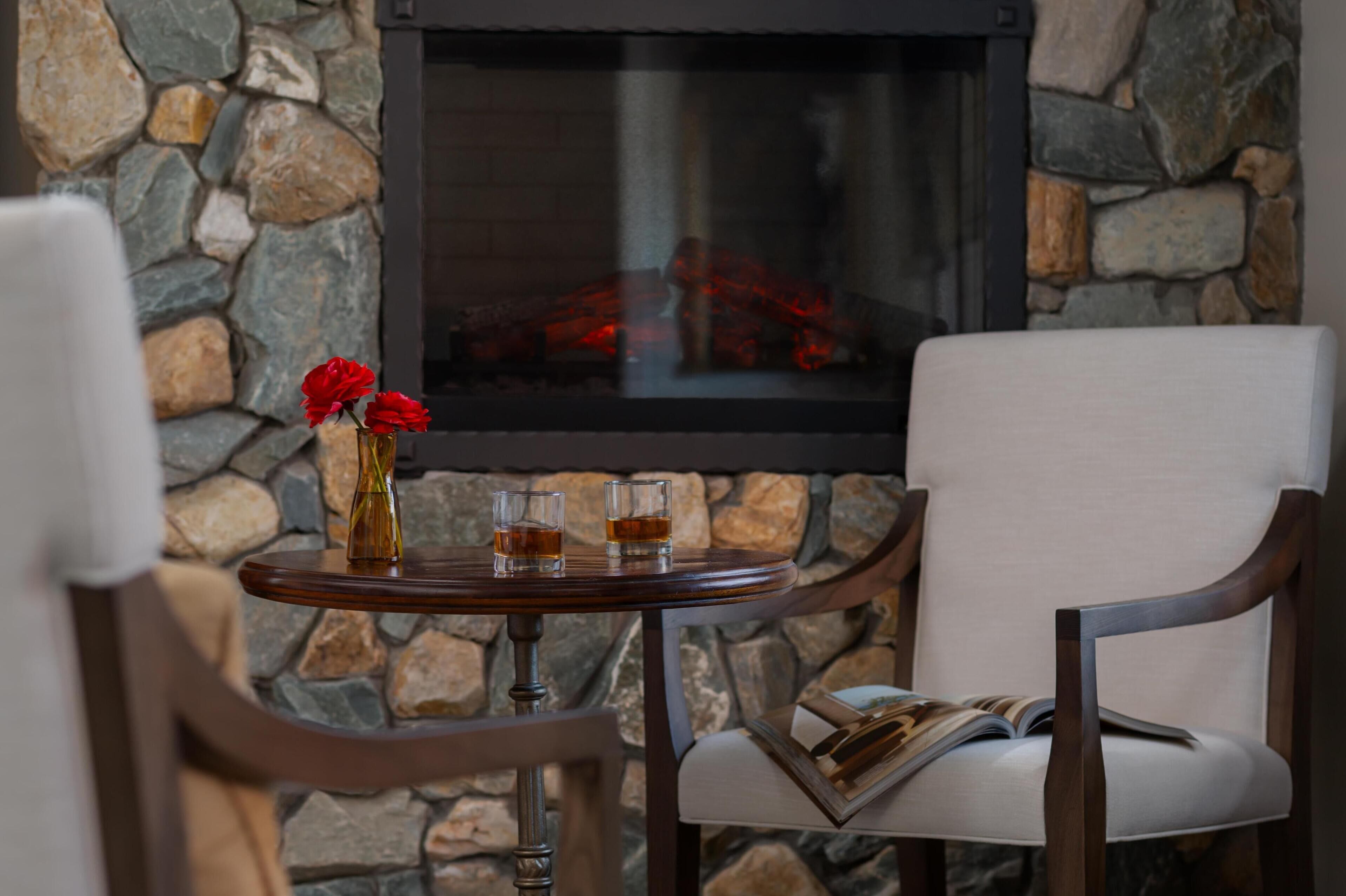 Un mur en pierre avec une cheminée, une table ronde en bois sur laquelle reposent deux verres de whisky et un vase rempli de fleurs rouges, et une chaise blanche avec un magazine.