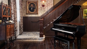 A grand piano, a staircase, and a wooden cabinet with a framed portrait in a room with patterned wallpaper and a wooden floor. - National Hotel Jackson, Tapestry Collection by Hilton (Jackson)