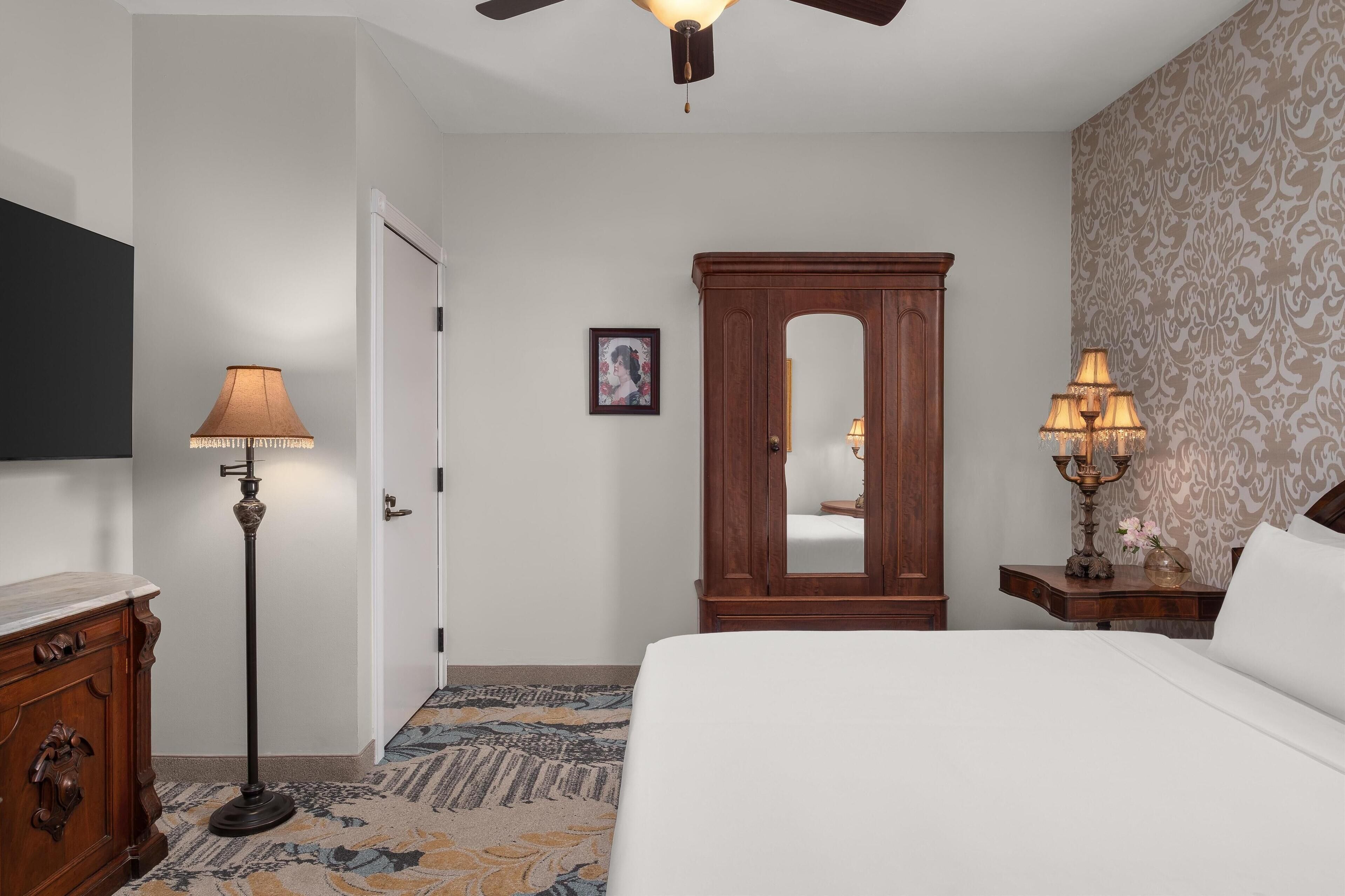 Una habitación de hotel con una cama, un armario de madera, una lámpara de pie, una mesita de noche con lámpara, un espejo y una alfombra estampada.