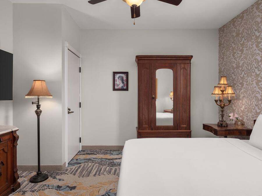 A hotel room with a bed, a wooden wardrobe, a floor lamp, a bedside table with a lamp, a mirror, and a patterned carpet.