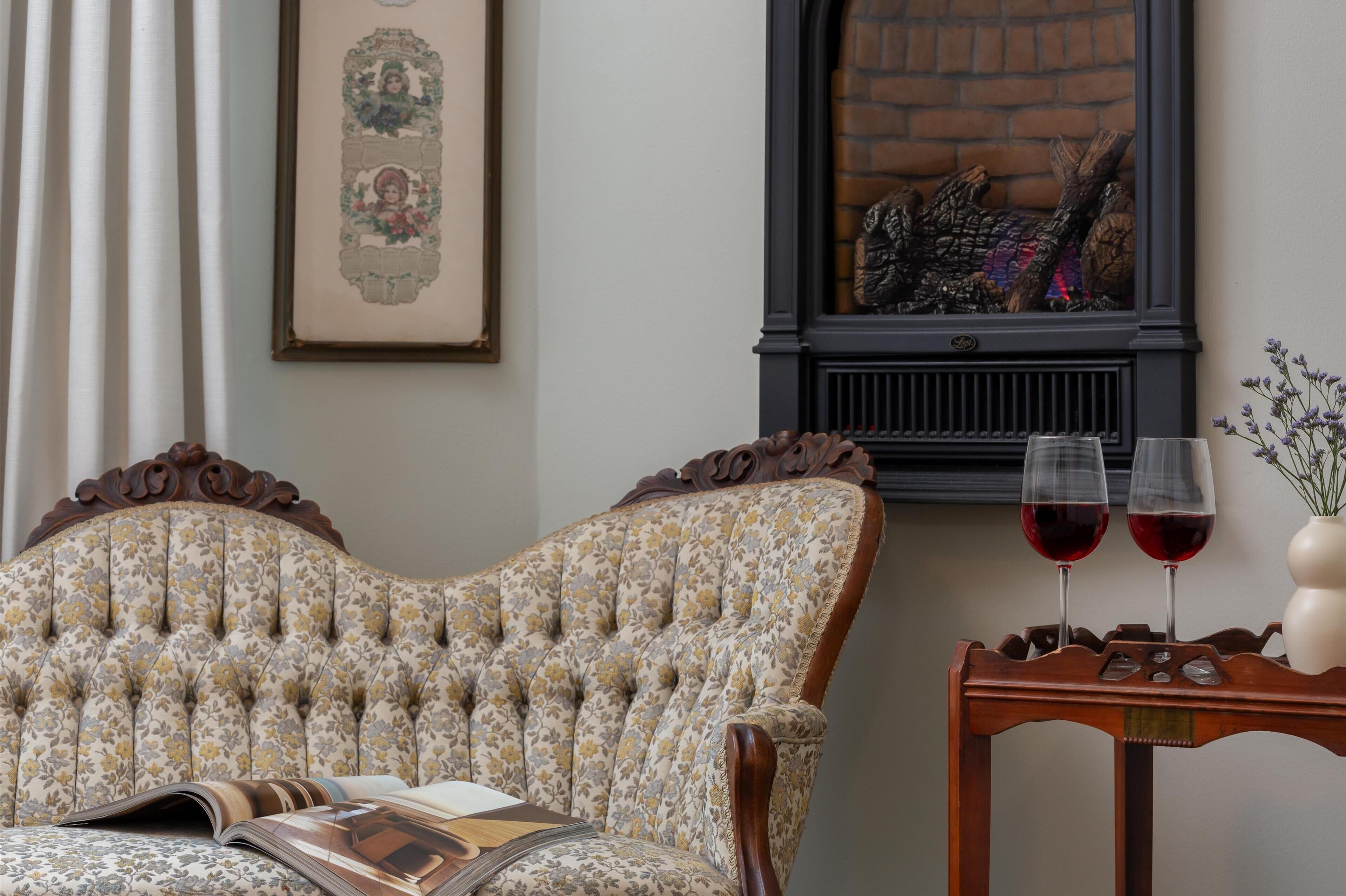 A room with a tufted upholstered chair, a framed artwork above a fireplace, and two glasses of red wine on a wooden side table.