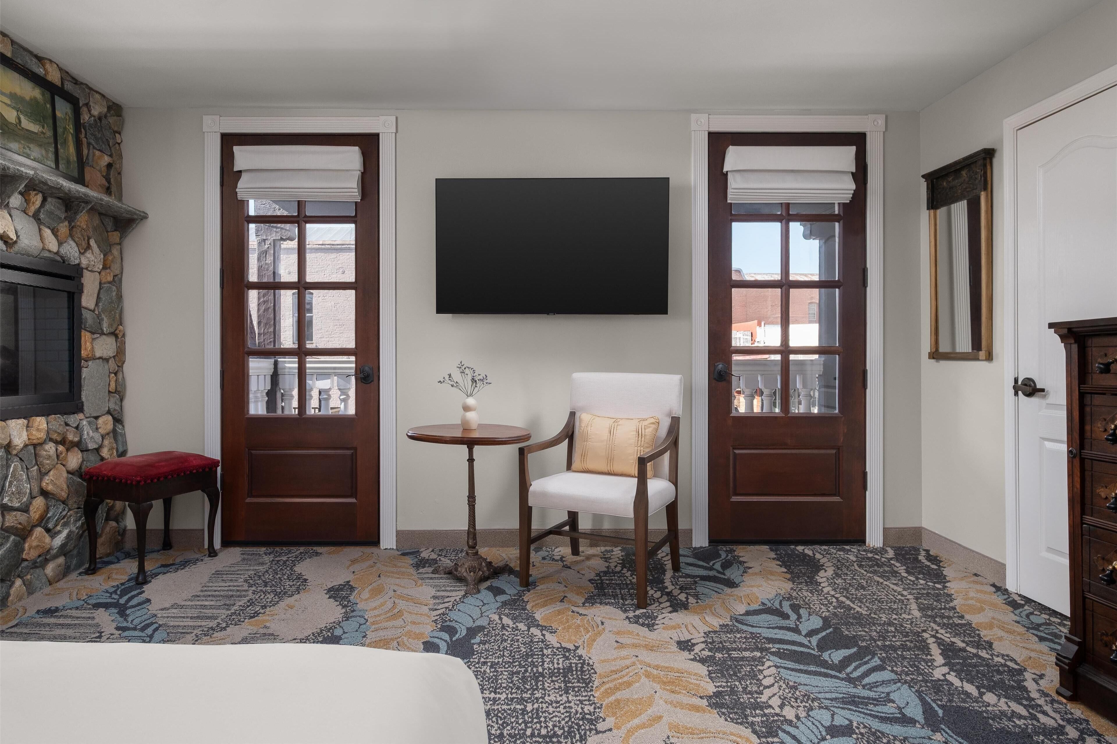 A hotel room with a stone fireplace, a flat-screen TV, a round table with a vase, a white armchair, and a patterned carpet.