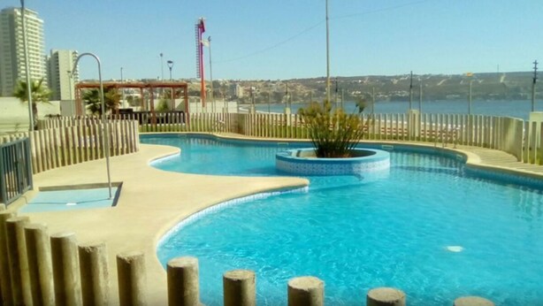 A swimming pool with a curved design, surrounded by a wooden fence and a view of the sea and buildings in the background.
