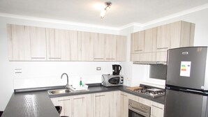 A modern kitchen with light wood cabinets, a stainless steel refrigerator, a microwave, a stove, and a sink.