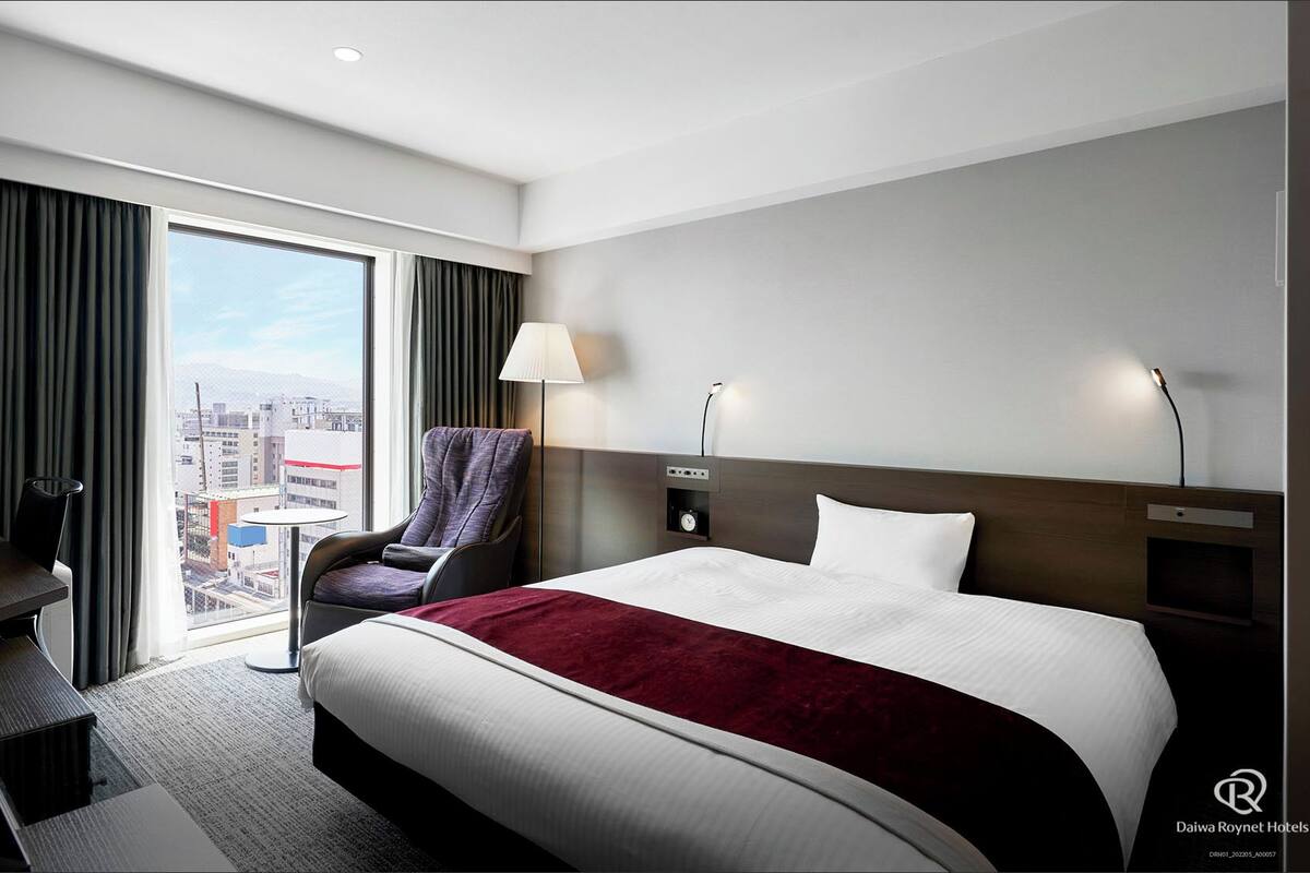 Een hotelkamer met een groot bed, een fauteuil, een bureau en uitzicht op de stad.