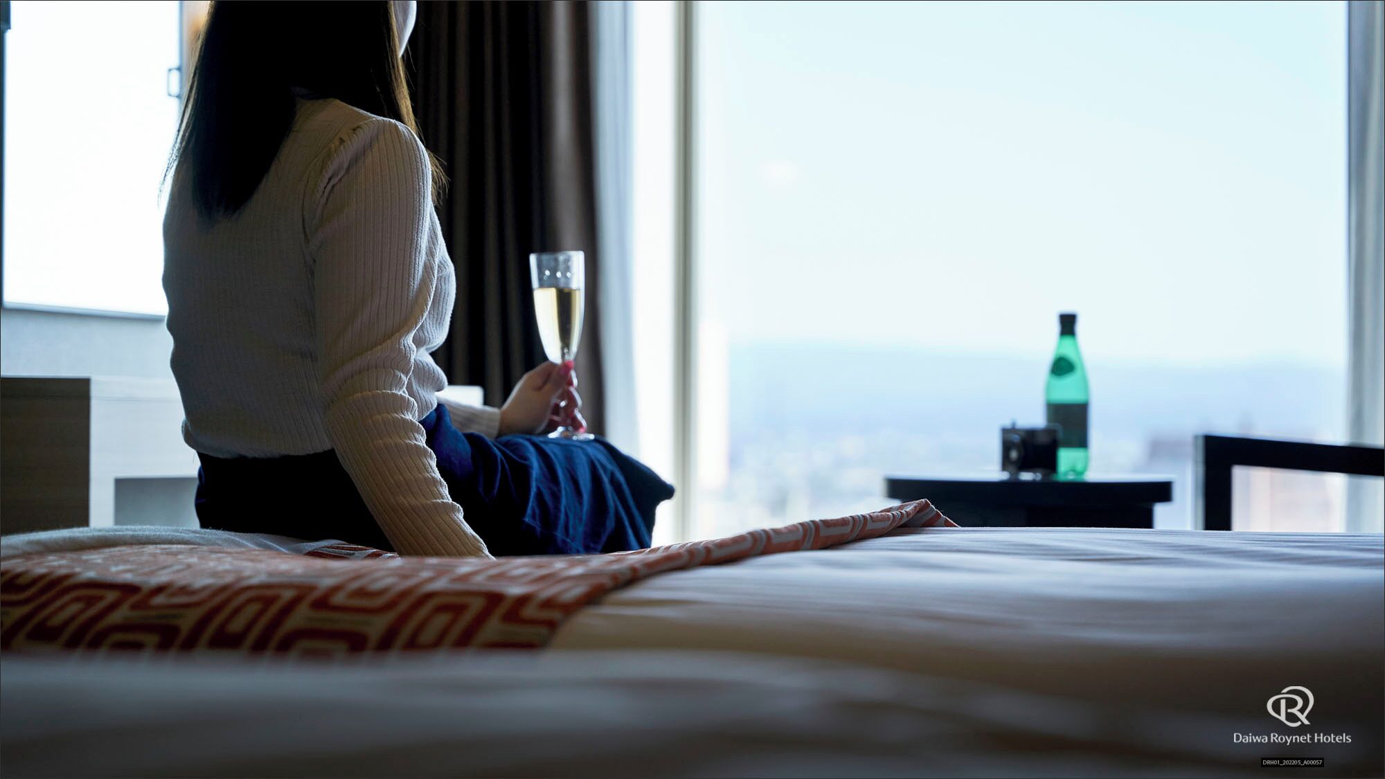 A hotel room with a bed, a glass of wine, a bottle, and a view of the city.