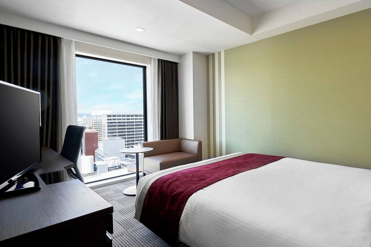 Een hotelkamer met een bed, een bureau met een computer, een stoel en een uitzicht op de stad door grote ramen.