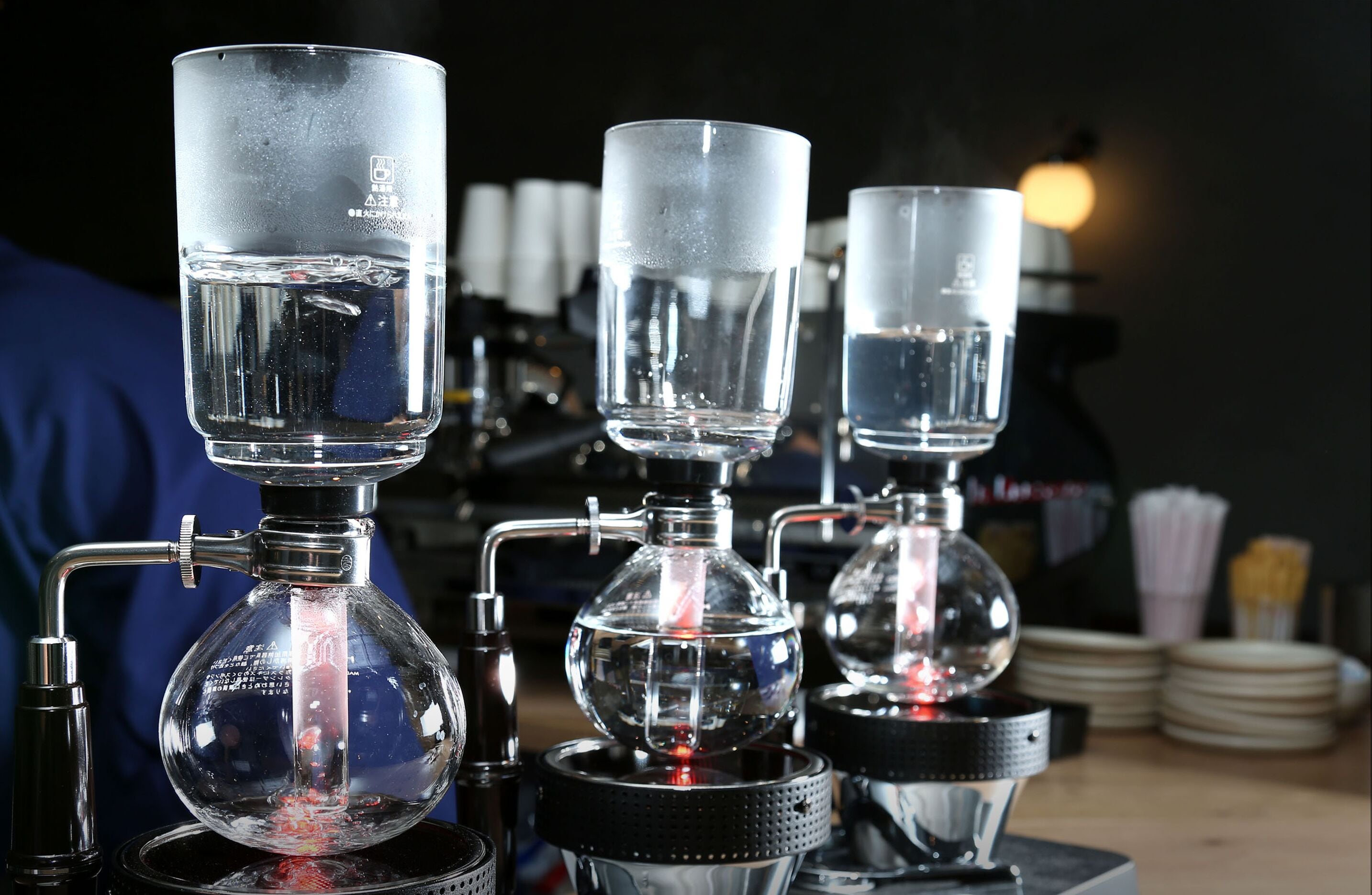 A coffee brewing setup with three transparent glass containers on a table, connected by metal pipes.