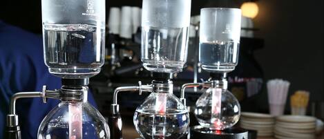 A coffee brewing setup with three transparent glass containers on a table, connected by metal pipes.