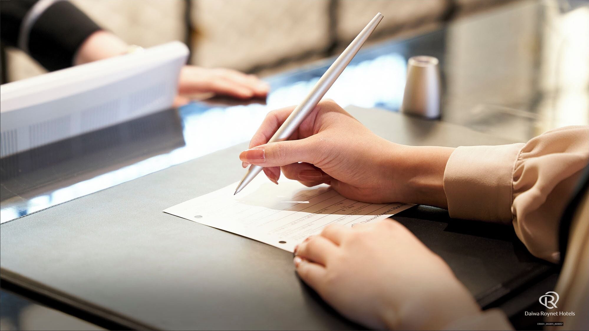 A person writing on a form at a table with a computer monitor in the background.