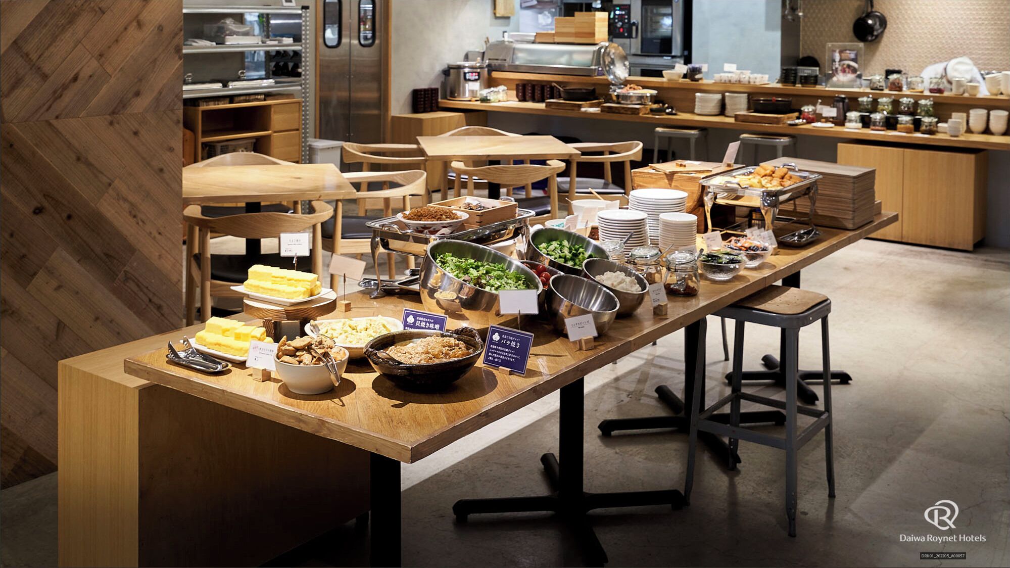 A buffet setup with various food items and utensils, wooden tables and chairs, and a kitchen area in the background.