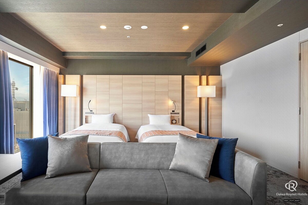 Een hotelkamer met een groot bed, twee nachtkastjes, een bank en uitzicht op de stad.