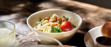 A bowl of salad with various vegetables and a tomato, a glass of white beverage, and a small container of butter on a wooden table.