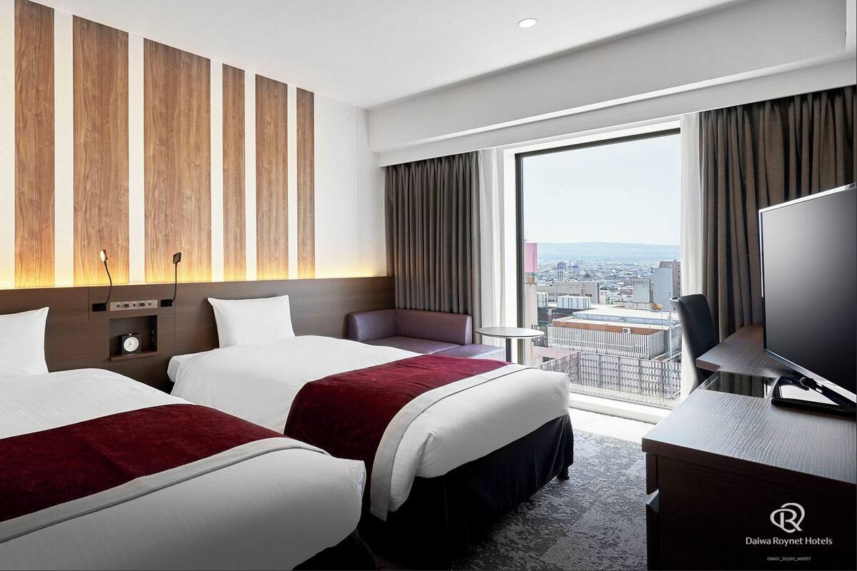 Een hotelkamer met twee bedden, een televisie, een bureau en een groot raam met uitzicht op de stad.