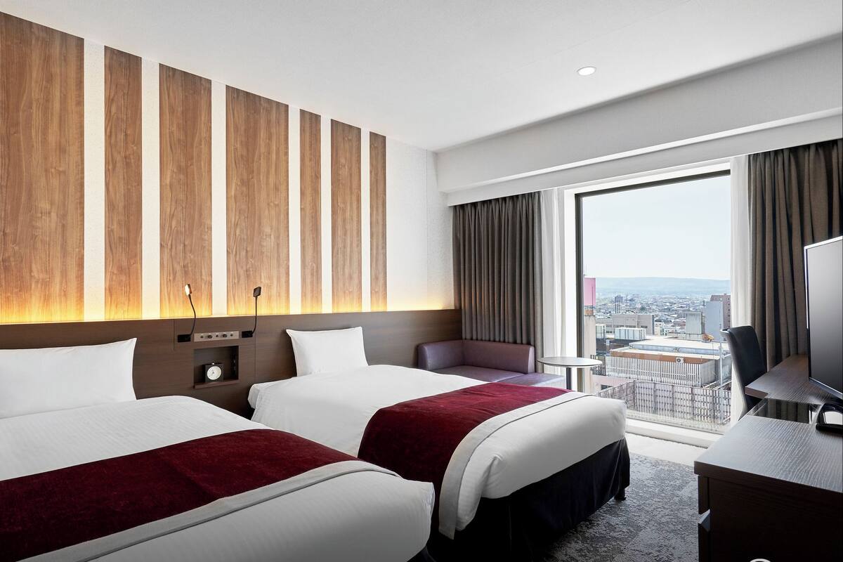 Hotelkamer met twee bedden, een bureau, een stoel en een groot raam met uitzicht op de stad.