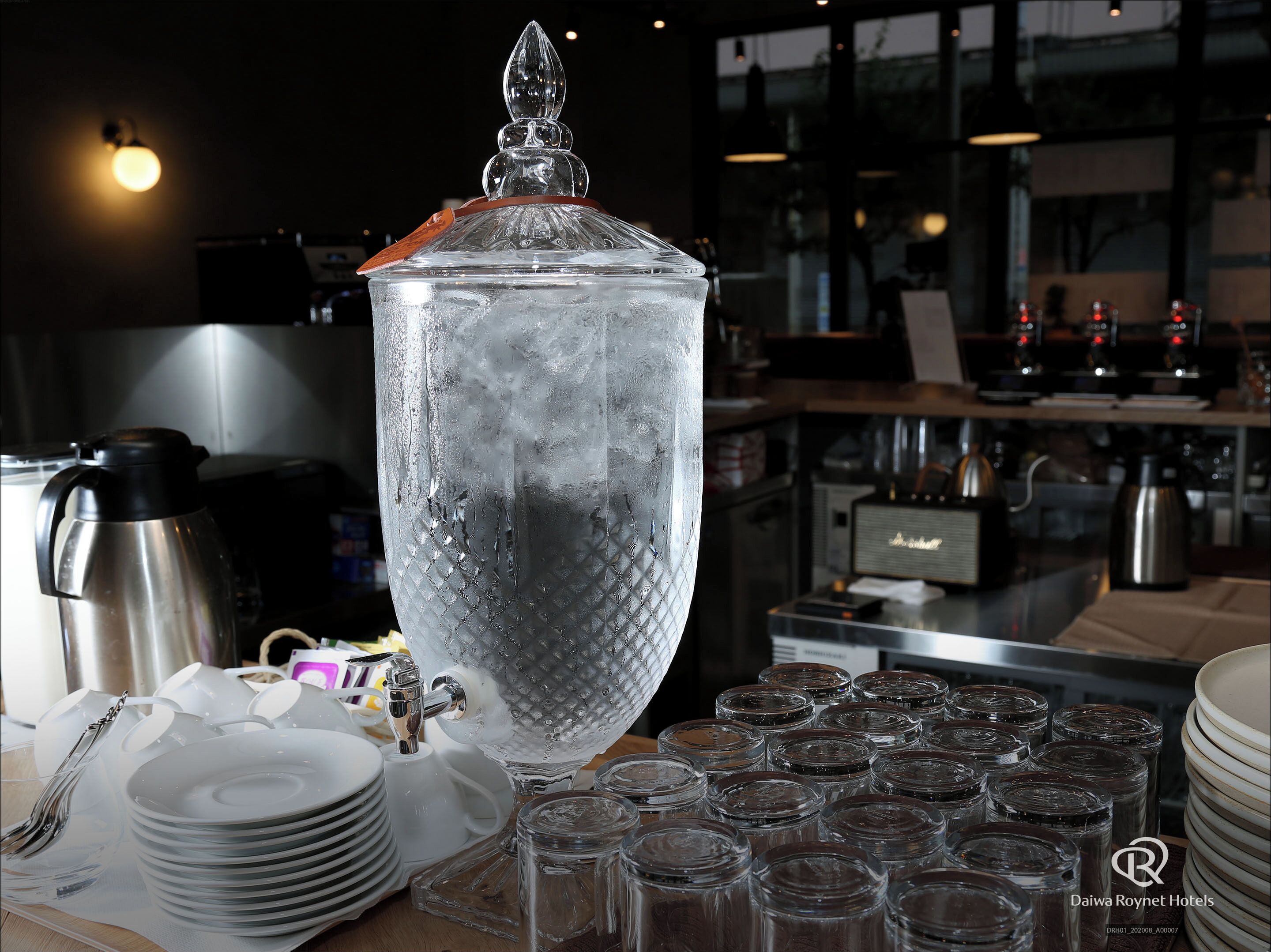 A large, clear glass container with a decorative lid, surrounded by stacked plates and small glass jars, likely in a restaurant or café setting.