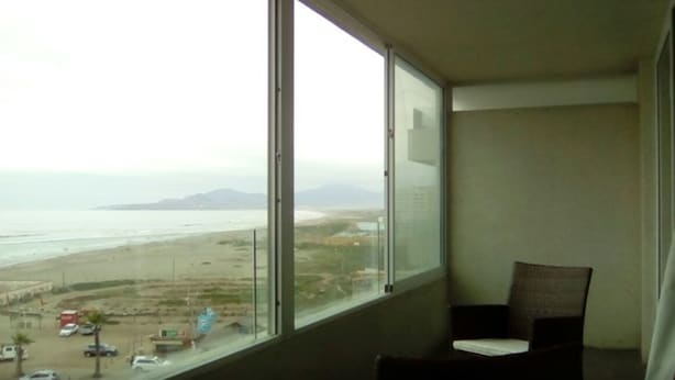 A room with a large window offering a view of a beach and distant mountains. - 47.- HNA Mirador El Faro (La Serena)