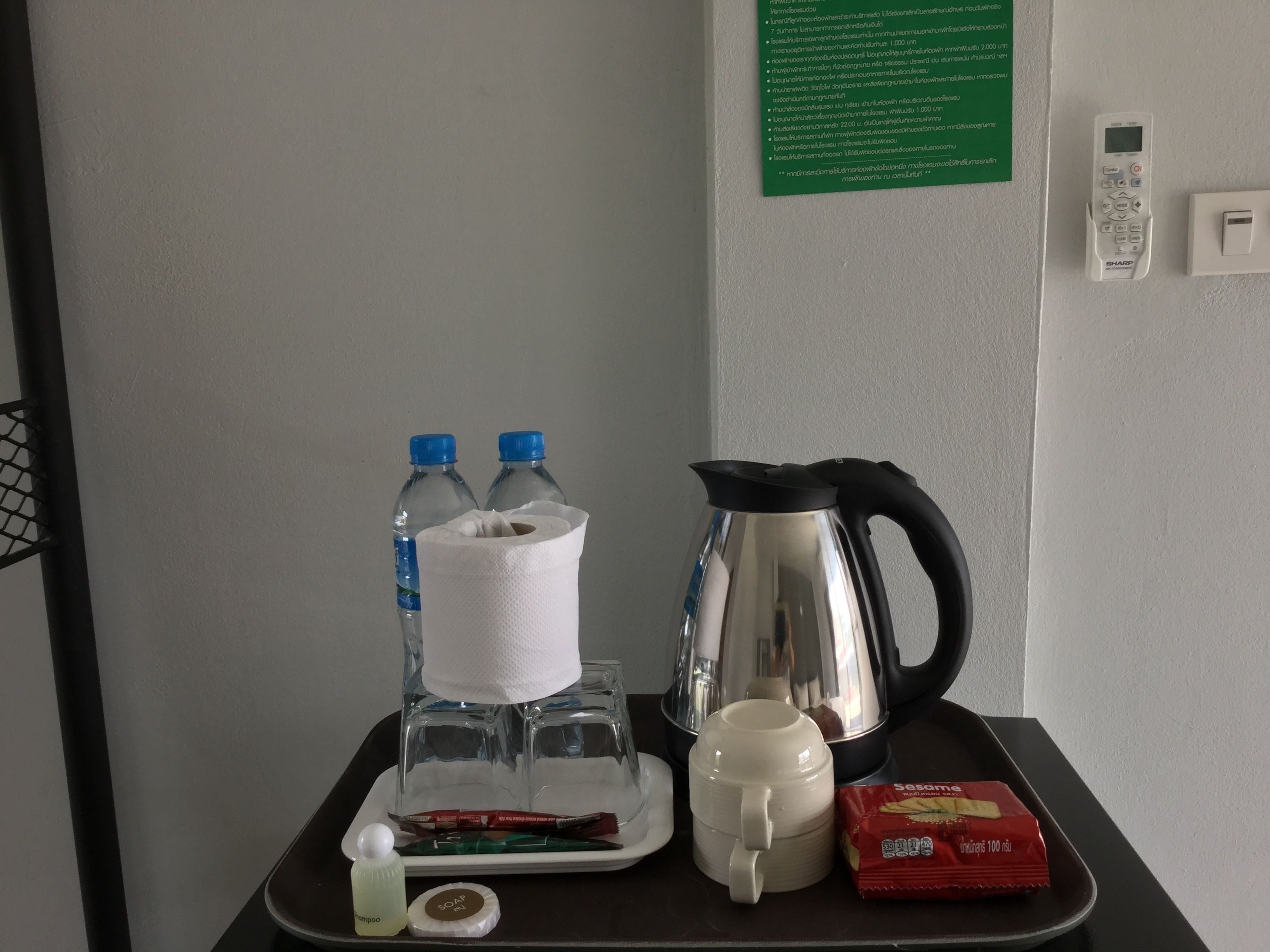 A hotel room's minibar with bottled water, a tissue roll, a kettle, a mug, shampoo, soap, and a snack pack.