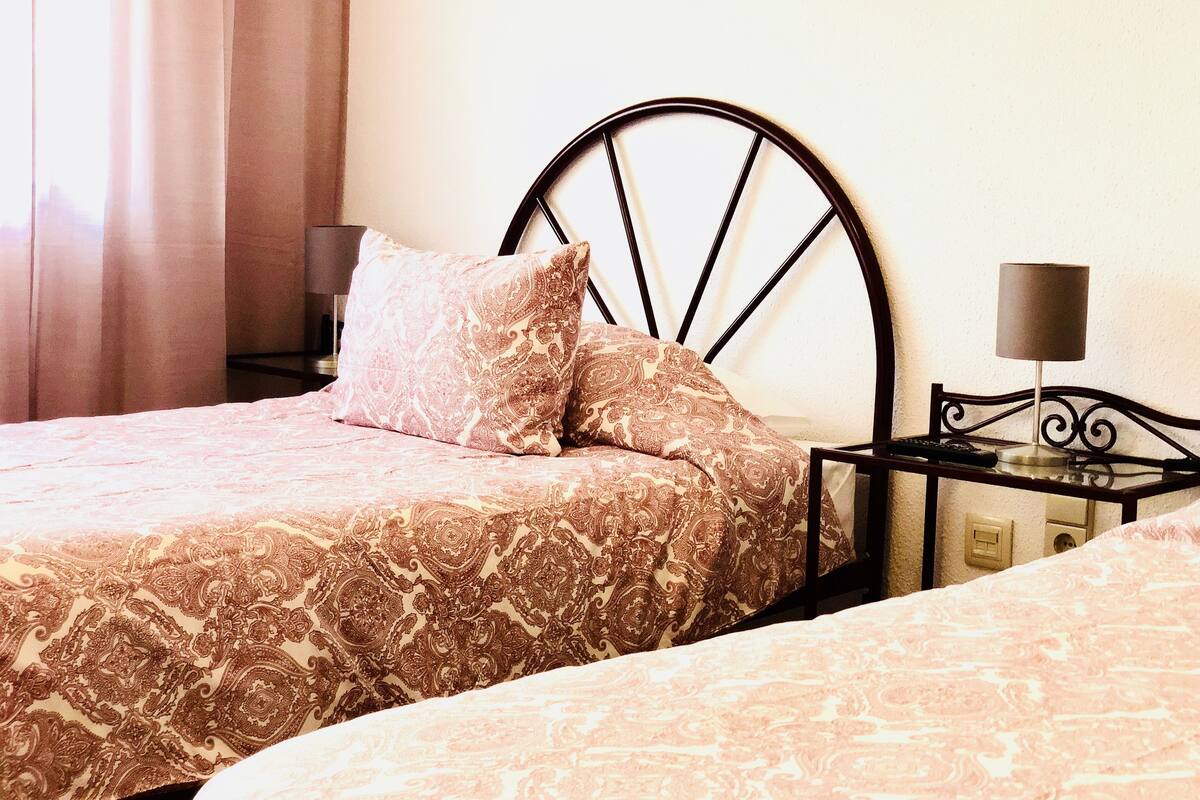 A double bed with a patterned bedspread and two pillows, a bedside table with a lamp, and a nightstand with a lamp.