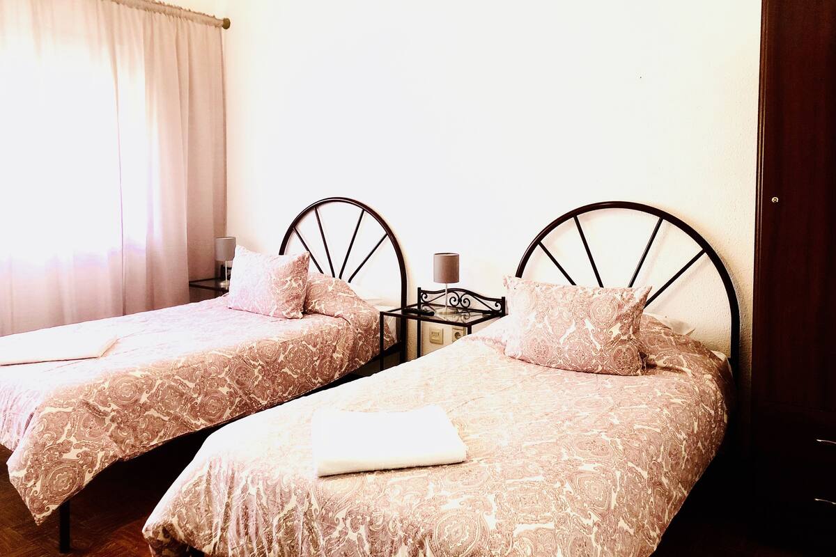 Two single beds with headboards, a nightstand with a lamp, and a window with curtains.